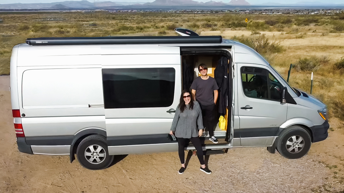 aerial drone view of man standing inside and a woman outside of the open sliding door of their silver Sprinter camper van. parked in an open land in Las Cruces, New Mexico.