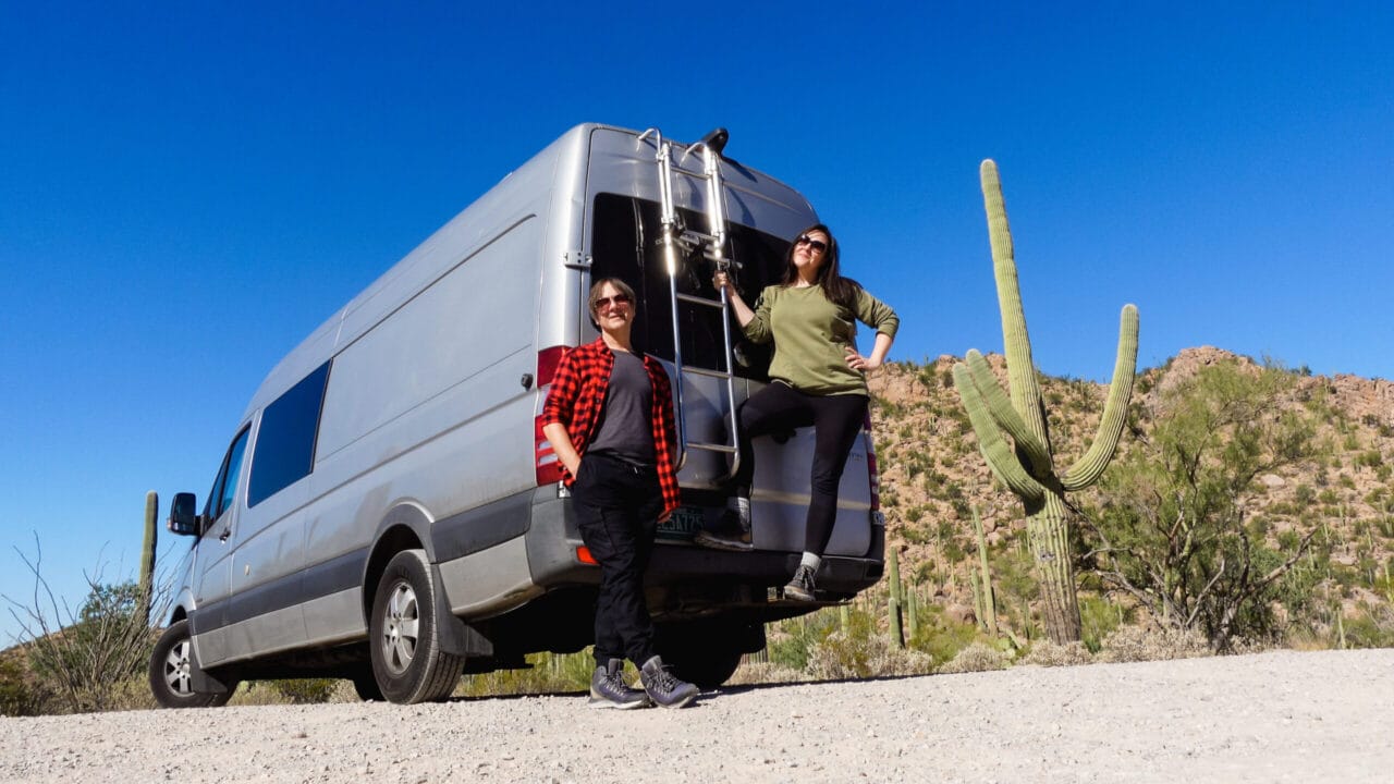 A man and woman pose with their silver Sprinter camper van in the Arizona desert, surrounded by cacti, while traveling the United States.